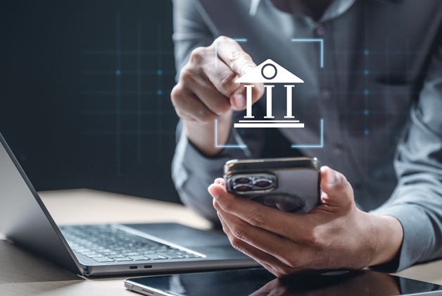 Mężczyzna w szarej koszuli używa smartfona, a na ekranie wyświetla się ikona banku. W tle widać laptopa.